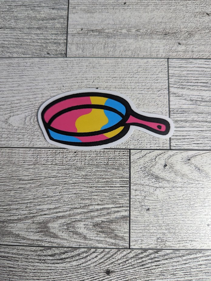 A sticker of a pan colored in the pan flag colors. The backdrop is a light wood grain.