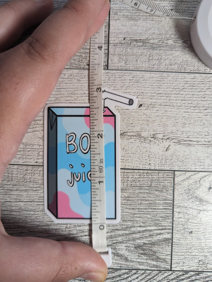 A juice box sticker with the words "BOY juice" on it. The colors are of the transmasculine flag. The backdrop is a light wood. A hand holds down a measuring tape which shows a height of approximately 3"