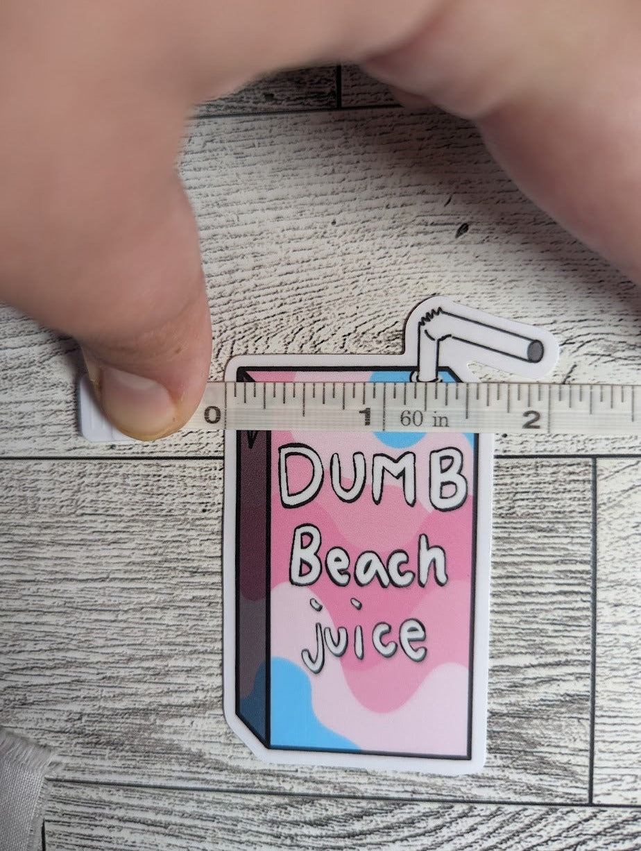 A juice box sticker with the words "DUMB Beach juice" on it. The colors are of the transfeminine flag. The backdrop is a light wood. A hand holds down a measuring tape which shows a width of approximately 2.165"