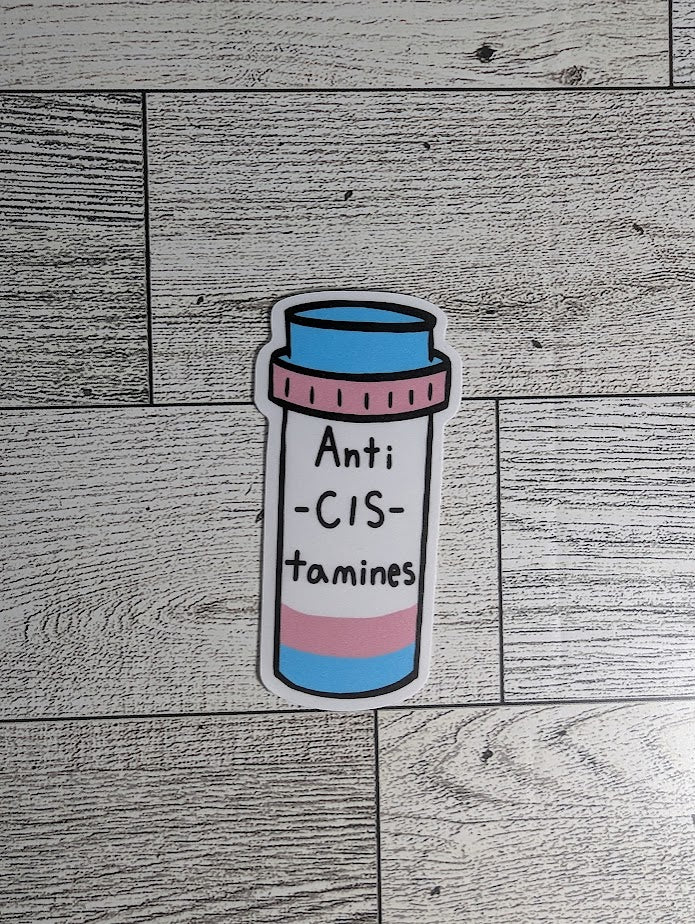 The sticker is of a pill bottle with the trans flag colors, light blue, light pink, and white. The words read "Anti-CIS-tamines." The backdrop is a light wood grain