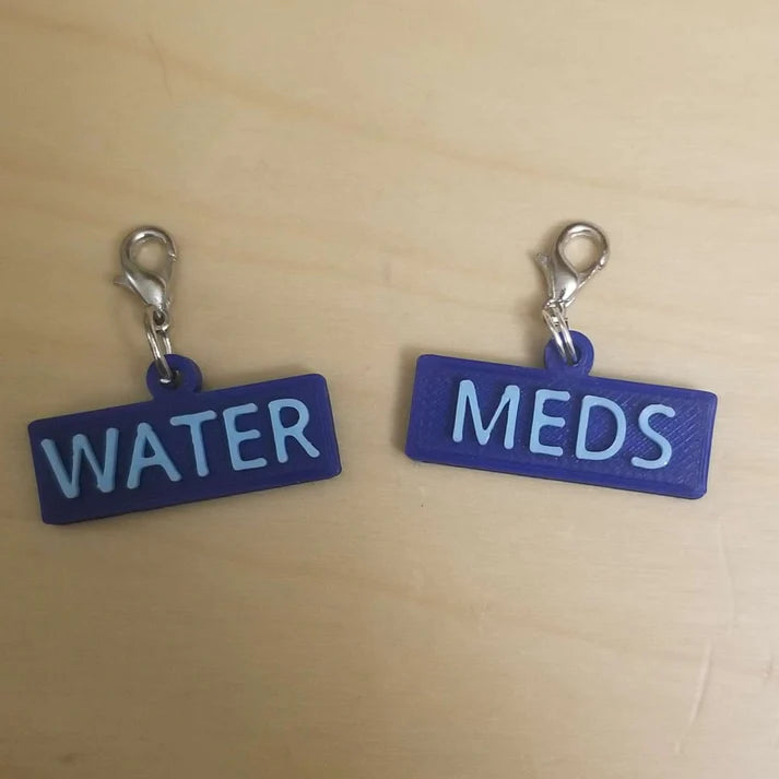 Two task charms with 3D printed light blue lettering on a dark blue background. A metal jump ring is used to connect the 3D printed piece to the clasp. The one on the left reads "water" and the other reads "meds"