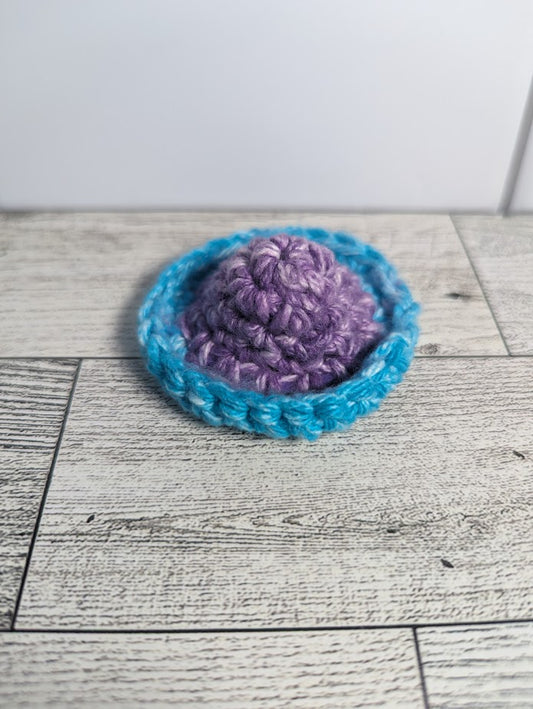 A crochet fidget with a purple center and a light blue rim. The shape of the fidget resembles a sombrero. The background is a light wood grain and a white tile.