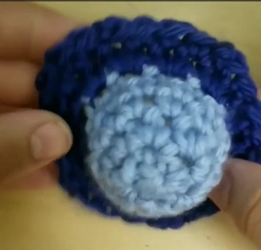 A crochet fidget is held against a light wood grain. The fidget mimics a pop-it in function. It has a light blue center and a dark blue skirt
