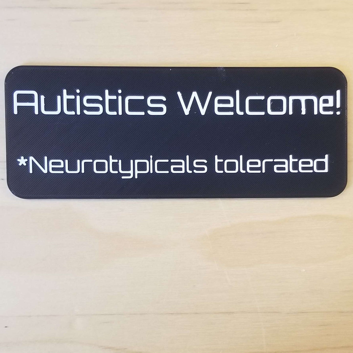 The sign reads "Autistics Welcome! *Neurotypicals tolerated" and the text is 3D printed in white. The background of the sign is a black rectangle with rounded corners. The sign is on a light tan wood desk.