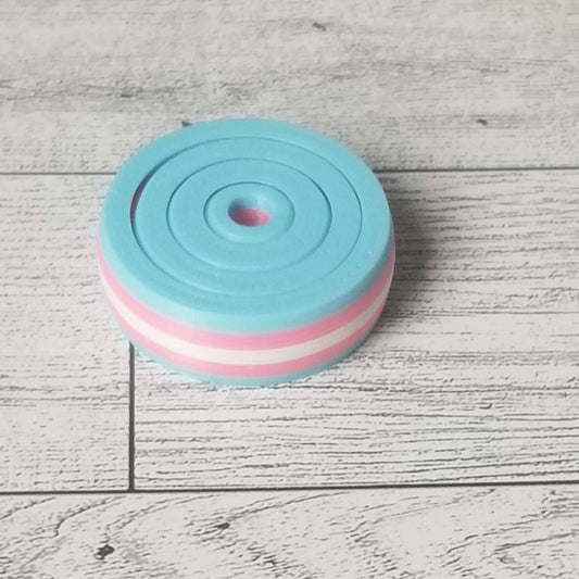 A small gyro fidget with colors striped light blue, light pink, white, light pink, and blue to mimic the trans flag. The background is a light woodgrain.