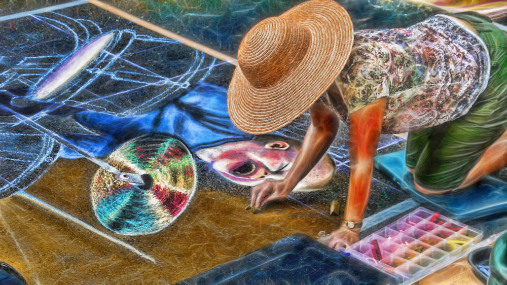Art sourced from Creative Commons. The art is of an artist chalking the sidewalk. The sidewalk has chalk drawings of drums and the artist has a medium skin tone wearing a sun hat, a T-shirt, and green shorts. The artist is kneeling on a knee pad and drawing the chalk art. A set of colorful chalk is next to the artist. 