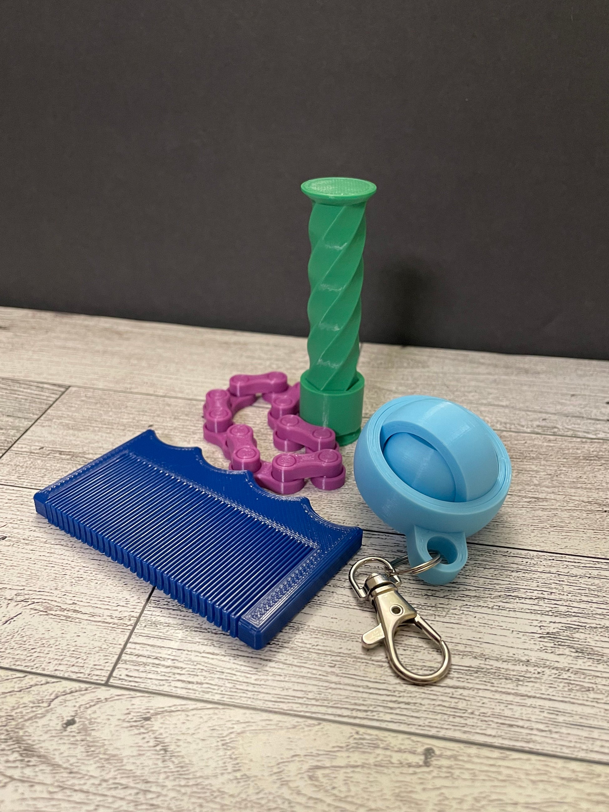 An image of a blue sensory comb, light blue small gyro fidget, purple medium chain fidget, and green spiral tower fidget. These are arranged on a light woodgrain table with a black backdrop.