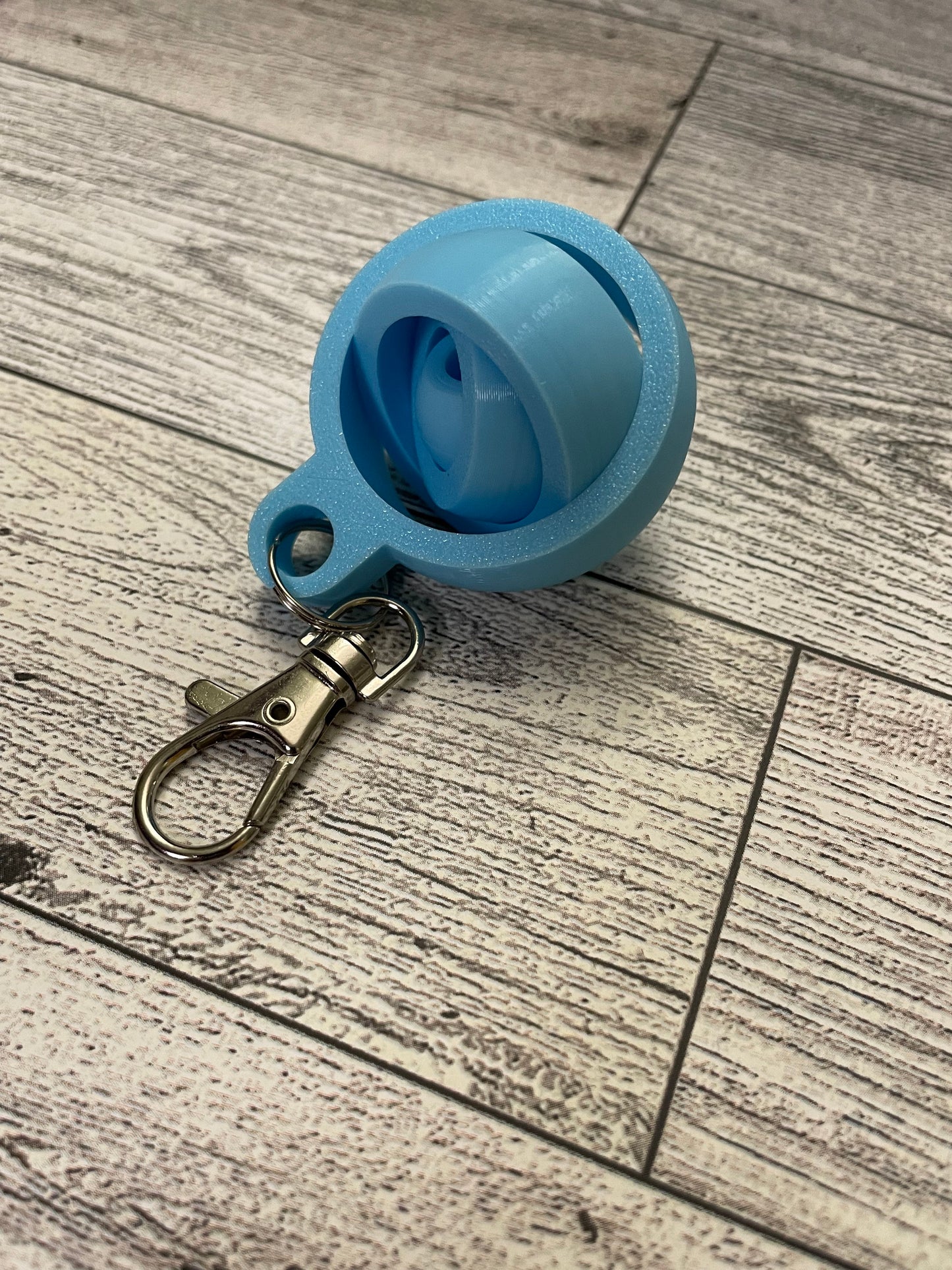 A small 3D printed gyro fidget that is about 1.5 inches in diameter on a wood background. It has four nested circles that can be spun and twirled. It is printed in light blue filament. The loop has a metal keychain attachment