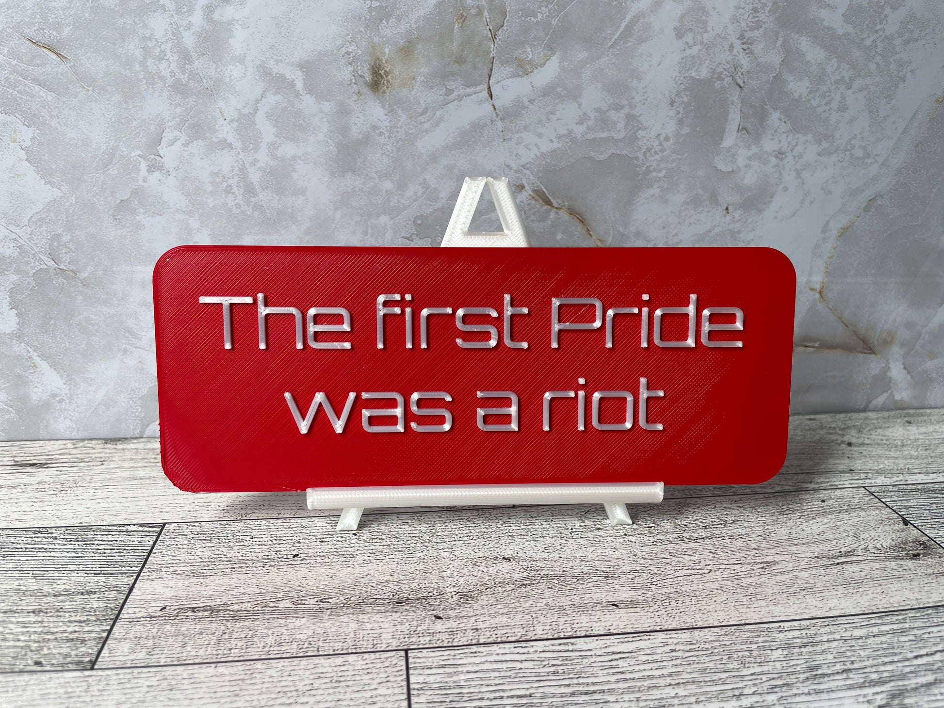 The text "The first Pride was a riot" is in shiny white text on a red sign. The sign is on a shiny white easel on a light woodgrain desk. The wall is a light marble with shades of white and tan.