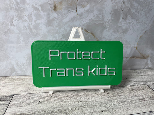 The text "Protect Trans kids" is in shiny white text on a green sign. The sign is on a shiny white easel on a light woodgrain desk. The wall is a light marble with shades of white and tan.