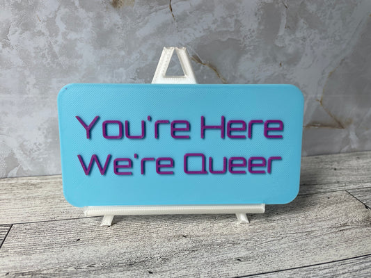 The text "You're Here We're Queer" is in purple text on a light blue sign. The sign is on a shiny white easel on a light woodgrain desk. The wall is a light marble with shades of white and tan.