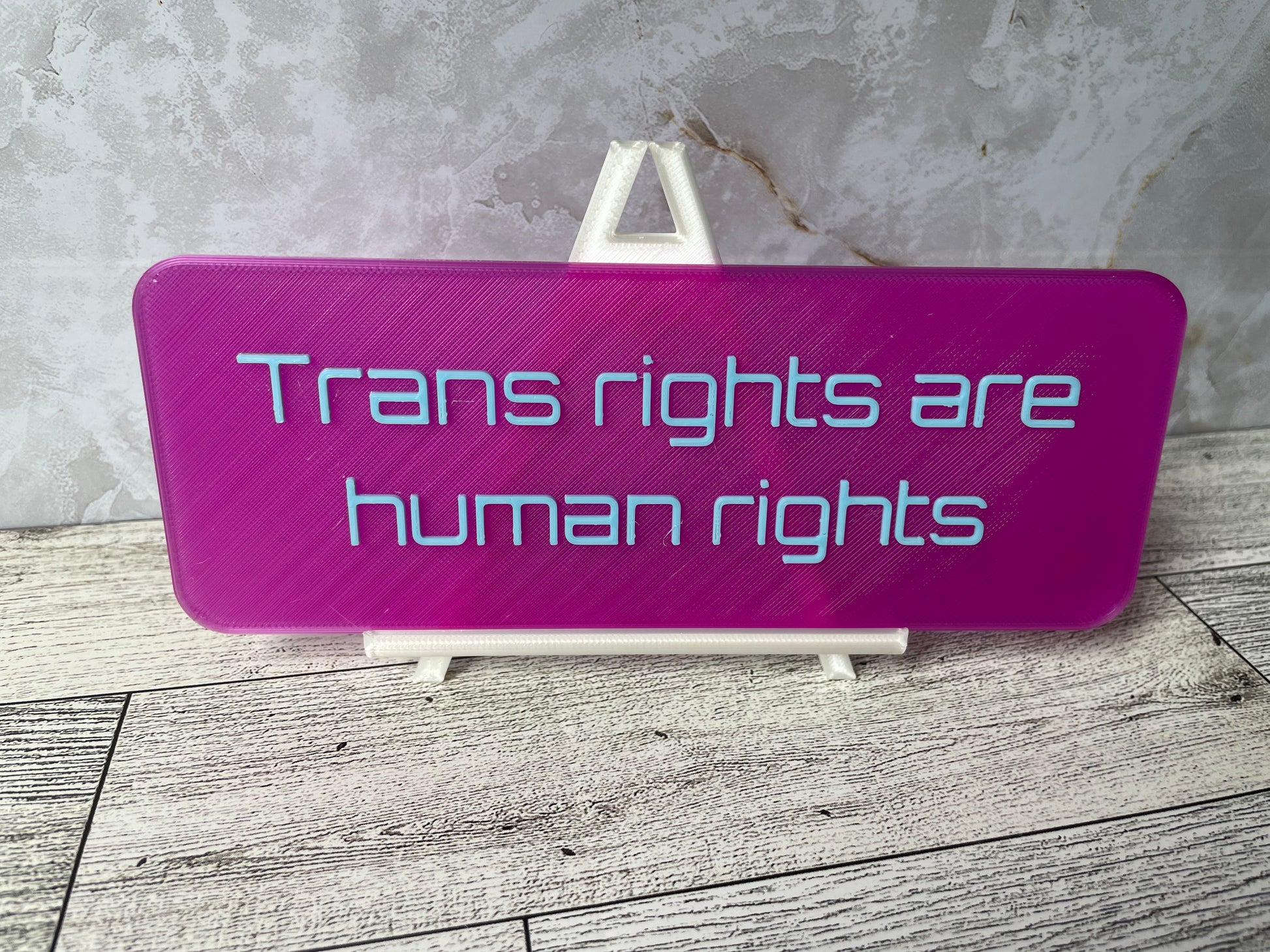 The text "Trans rights are human rights" is in light blue text on a translucent purple sign. The sign is on a shiny white easel on a light woodgrain desk. The wall is a light marble with shades of white and tan.