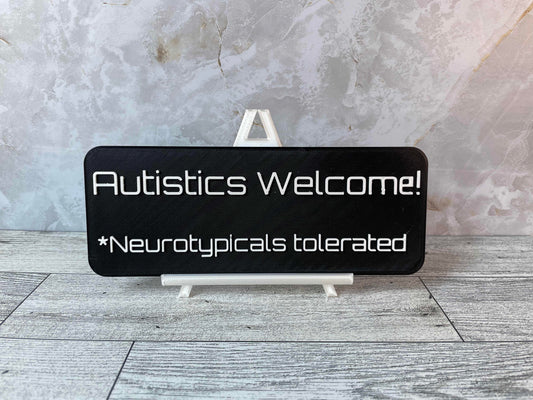 The text "Autistics Welcome! *Neurotypicals tolerated" is in white text on a black sign. The sign is on a shiny white easel on a light woodgrain desk. The wall is a light marble with shades of white and tan.