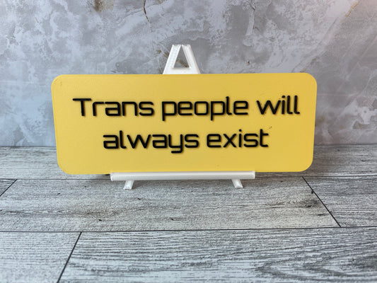 The text "Trans people will always exist" is in black text on a yellow sign. The sign is on a shiny white easel on a light woodgrain desk. The wall is a light marble with shades of white and tan.