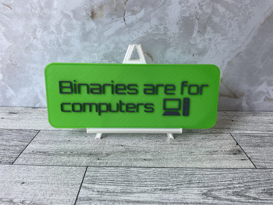 The text "Binaries are for computers" is in gray text on a watermellon-lime green sign. An old desktop computer and monitor icon is also on the sign.The sign is on a shiny white easel on a light woodgrain desk. The wall is a light marble with shades of white and tan.