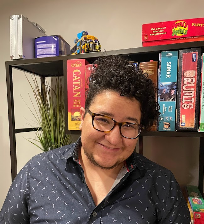 A photo of Ash who has olive skin, dark brown curly hair and brown eyes. Their hair is cut in a loose afro with an undercut on the side. They are wearing tortoise colored glasses and a dark blue button-up with silver birds on it. The background has a bookshelf with board games on it such as Catan and a fake plant. The wall is a light off-white color.
