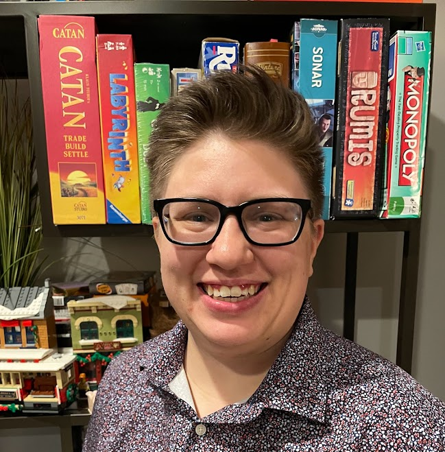 A photo of Andy has a light skin color typical of caucasians, a light brown hair color and blue eyes. They have a short haircut that flips to the side. They are wearing black glasses and a spotted blue, red, white button-up.  The background has a bookshelf with a Lego set and board games on it such as Catan and a fake plant. The wall is a light off-white color.