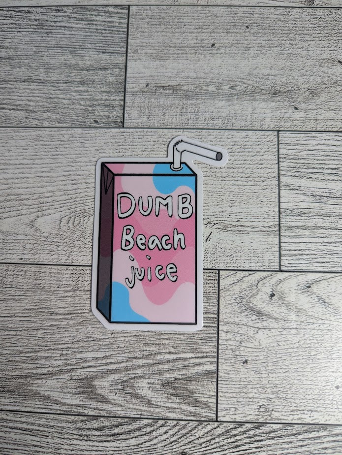 A juice box sticker with the words "DUMB Beach juice" on it. The colors are of the transfeminine flag. The backdrop is a light wood.