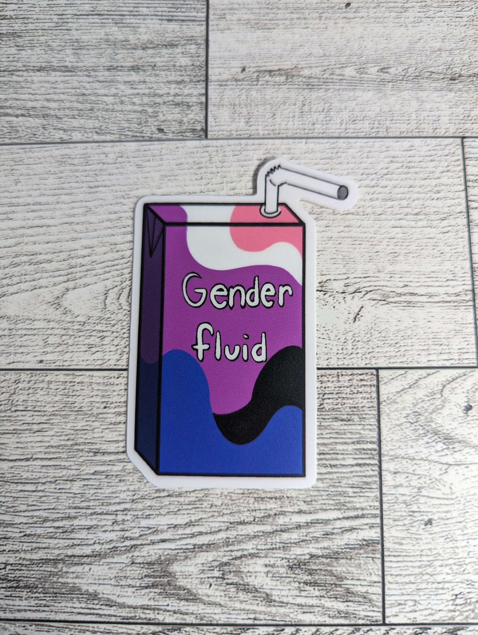 A juice box sticker with the words "Gender fluid" on it. The colors are of the genderfluid flag. The backdrop is a light wood.