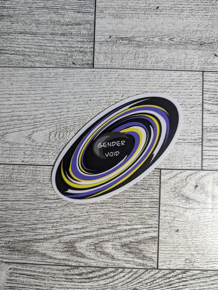 A sticker with the words "GENDER VOID" at the center of a black hole with a nonbinary flag colors. The backdrop is a light wood grain.