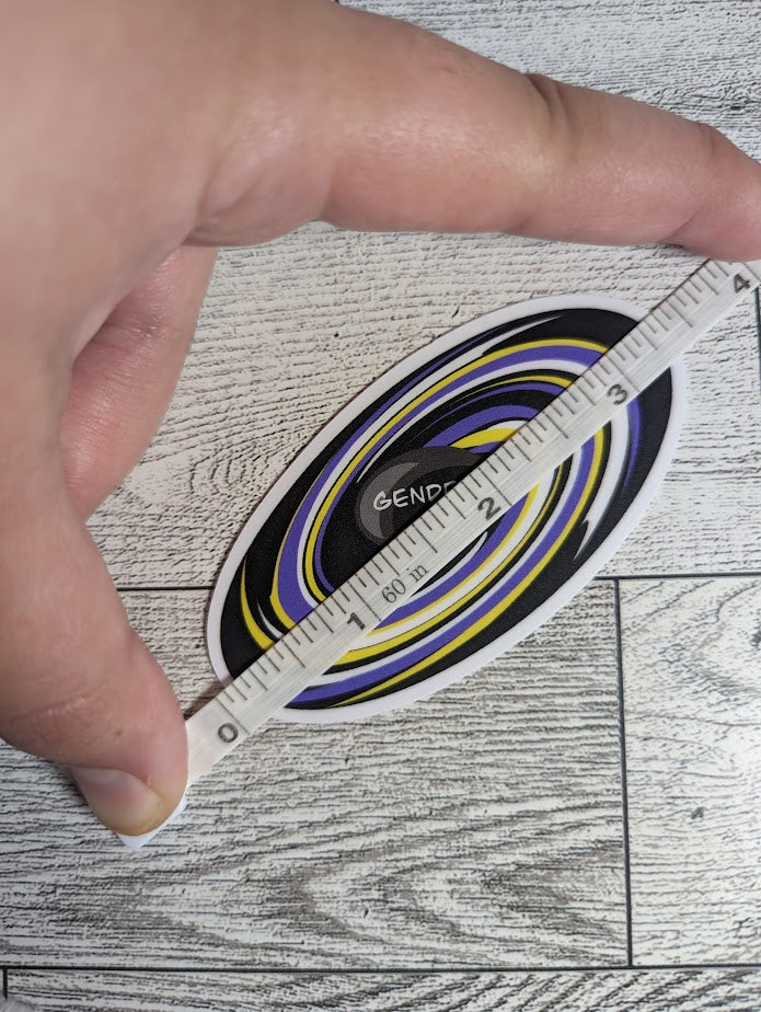 A sticker with the words "GENDER VOID" at the center of a black hole with a nonbinary flag colors. The backdrop is a light wood grain. A hand holds a measuring tape to show the diagonal.