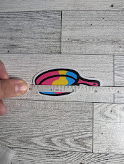 A sticker of a pan colored in the pan flag colors. The backdrop is a light wood grain. A hand holds a measuring tape to show the width.