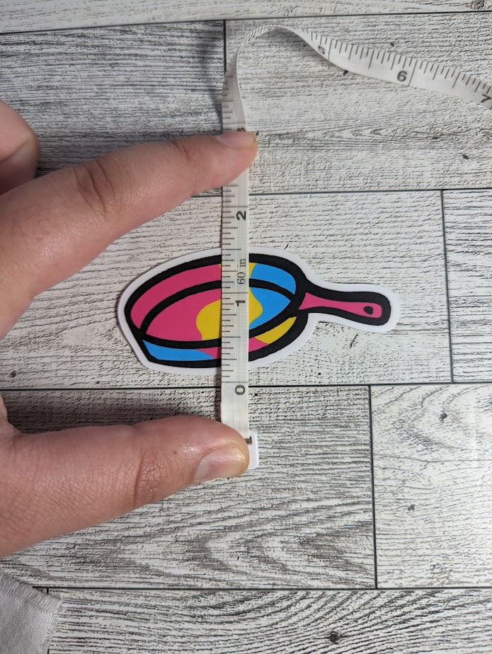 A sticker of a pan colored in the pan flag colors. The backdrop is a light wood grain. A hand holds a measuring tape to show the height
