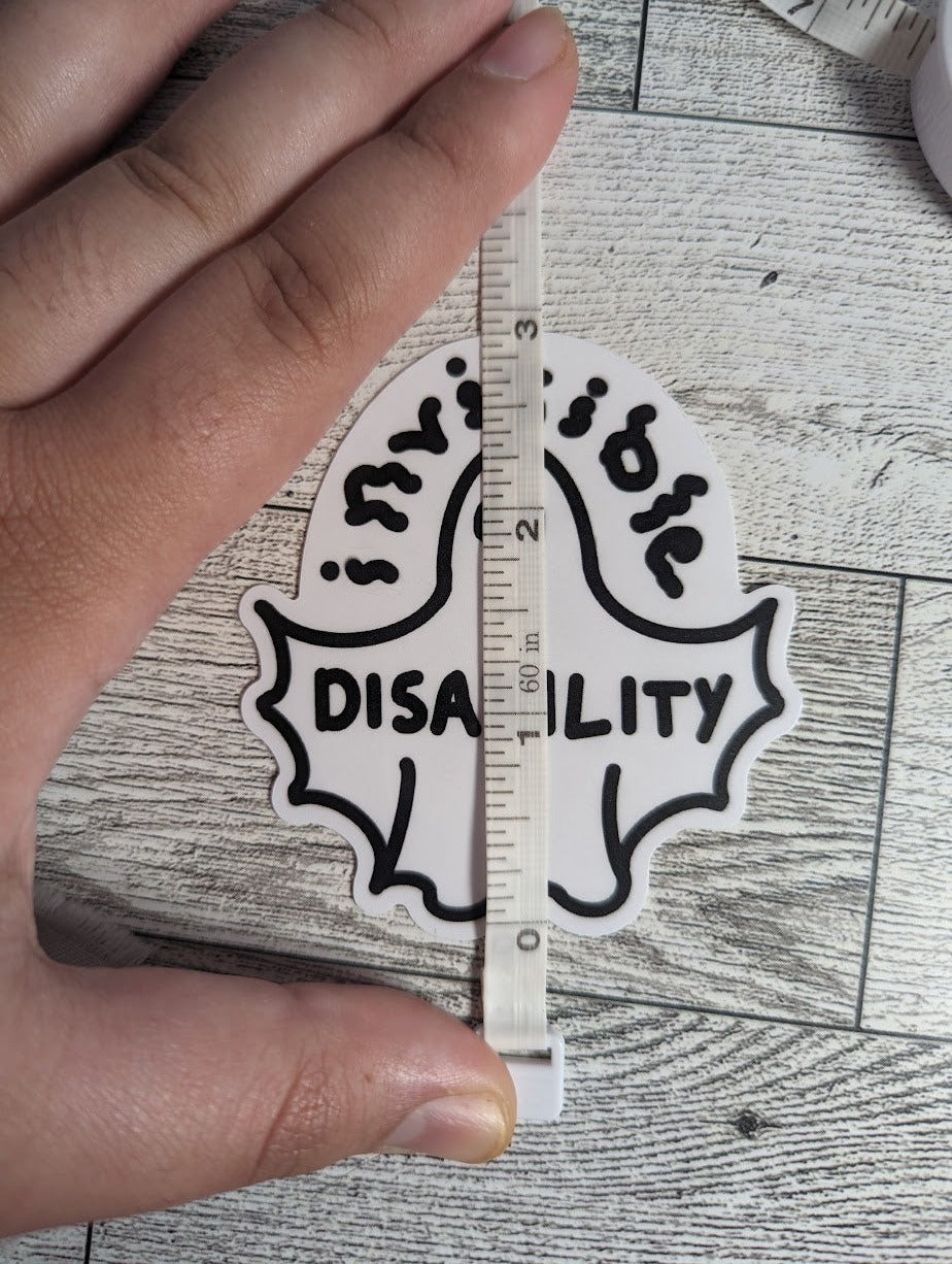 A ghost sticker with the word "invisible" drawn in squiggle font above and "DISABILITY" is across the wingspan. The ghost's arms are bat-like shape into the body. There is a light wood backdrop. A hand holds down a measuring tape to show a height of 3"