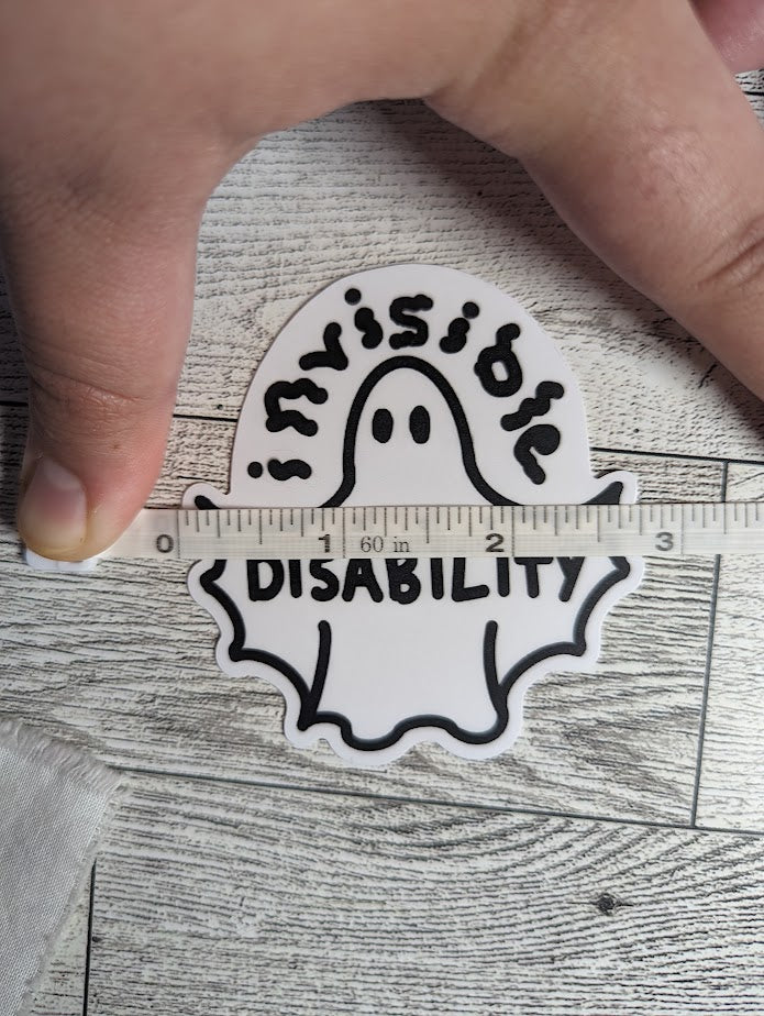 A ghost sticker with the word "invisible" drawn in squiggle font above and "DISABILITY" is across the wingspan. The ghost's arms are bat-like shape into the body. There is a light wood backdrop. A hand holds down a measuring tape to show a width of 2.75"
