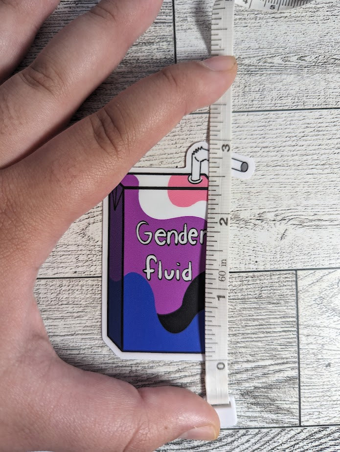 A juice box sticker with the words "Gender fluid" on it. The colors are of the genderfluid flag. The backdrop is a light wood. A hand is holding down a measuring tape that shows the height is approximately 3"