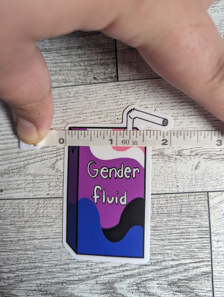 A juice box sticker with the words "Gender fluid" on it. The colors are of the genderfluid flag. The backdrop is a light wood. A hand is holding down a measuring tape that shows the width is approximately 2.165"