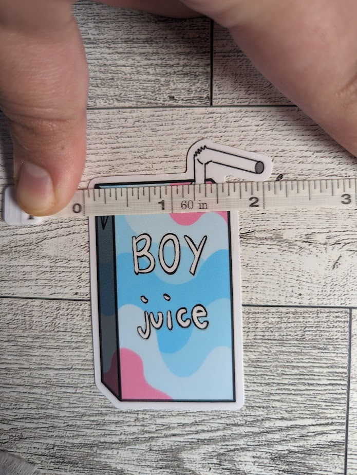 A juice box sticker with the words "BOY juice" on it. The colors are of the transmasculine flag. The backdrop is a light wood. A hand holds down a measuring tape which shows a width of approximately 2.165"