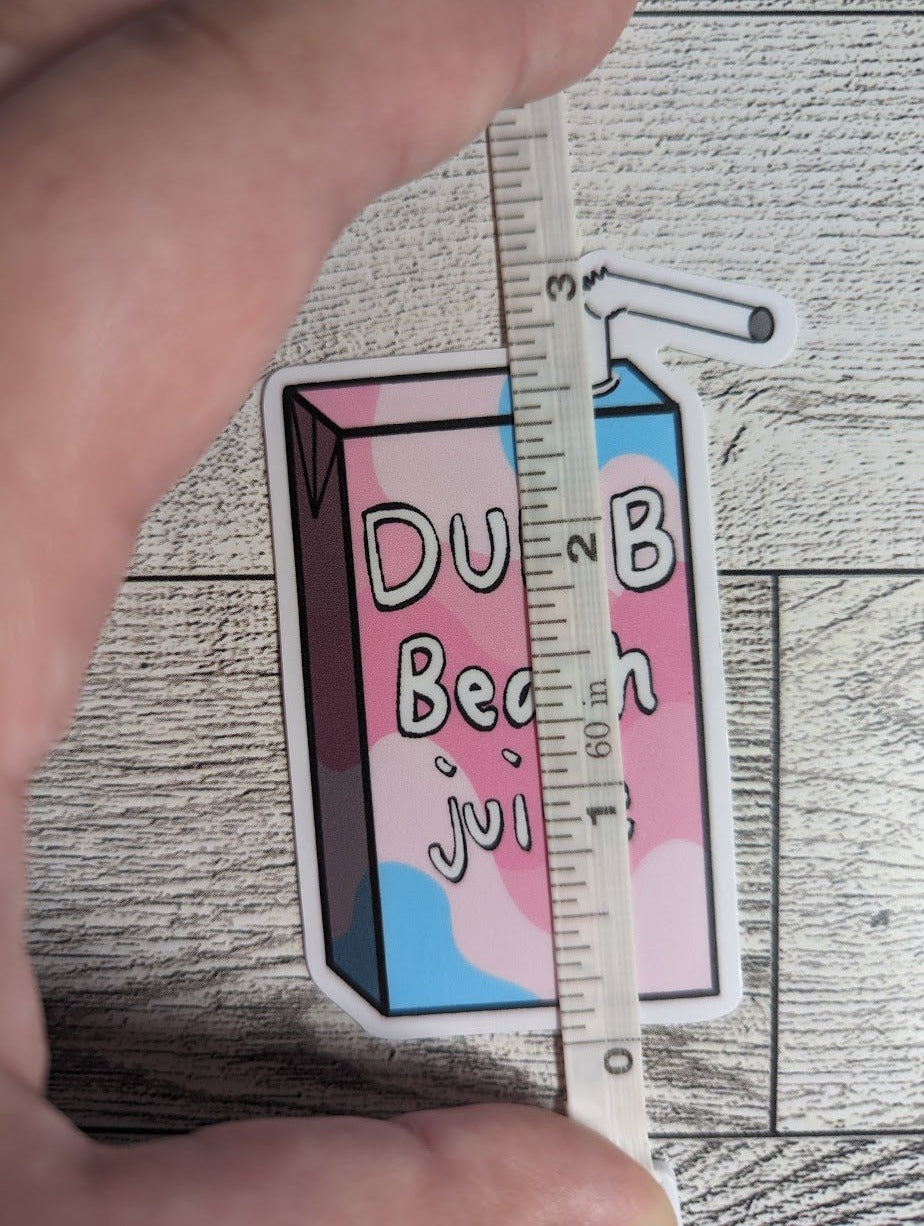 A juice box sticker with the words "DUMB Beach juice" on it. The colors are of the transfeminine flag. The backdrop is a light wood. A hand holds down a measuring tape which shows a height of approximately 3"
