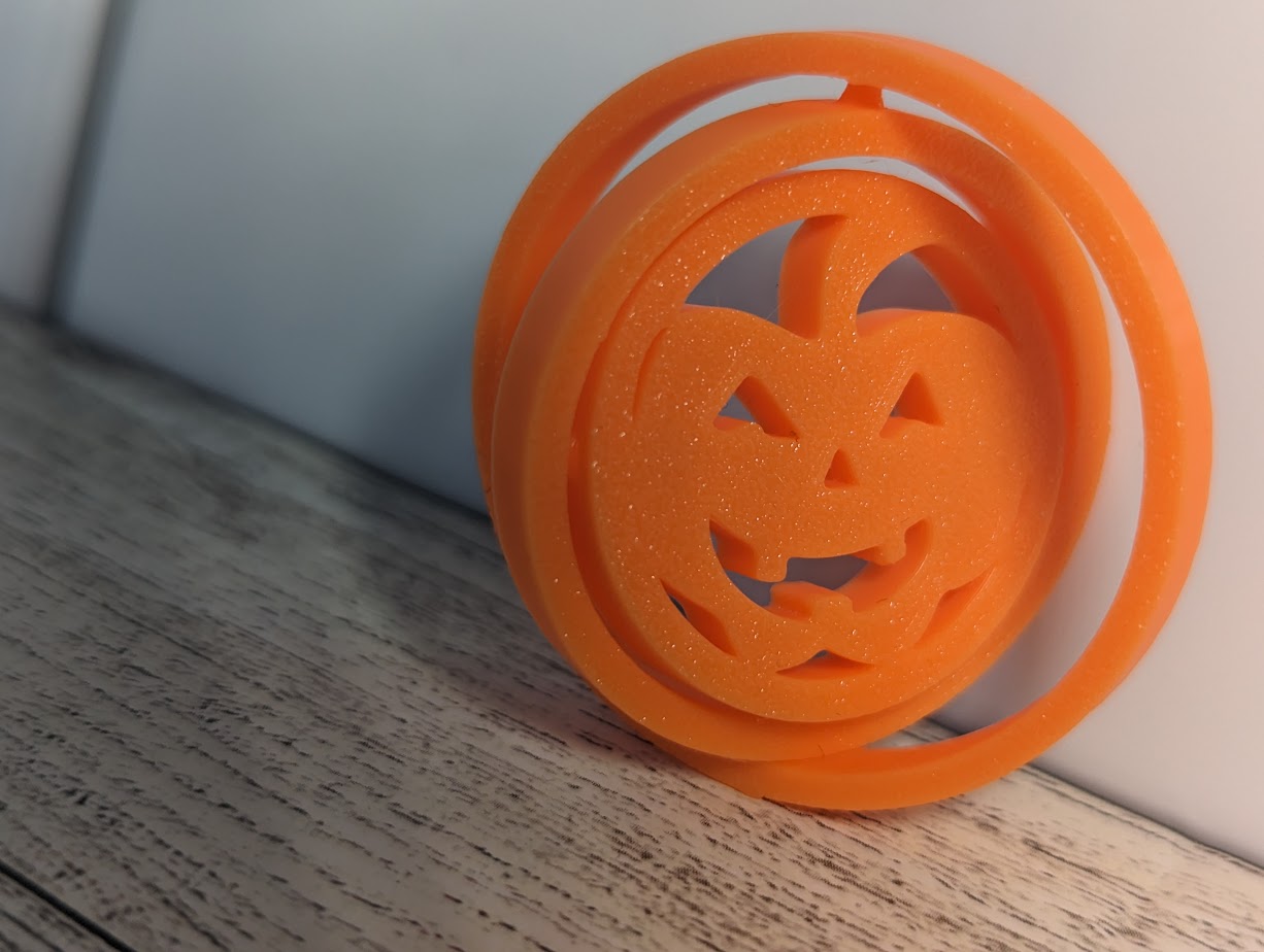 An orange circle jack-o-lantern fidget is spun partially and leaning against a white tile and light woodgrain background.