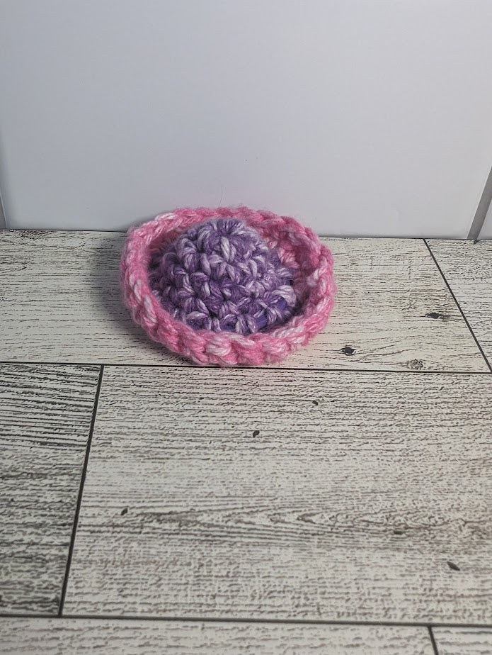 A crochet fidget with a purple center and a pink rim. The shape of the fidget resembles a sombrero. The background is a light wood grain and a white tile.
