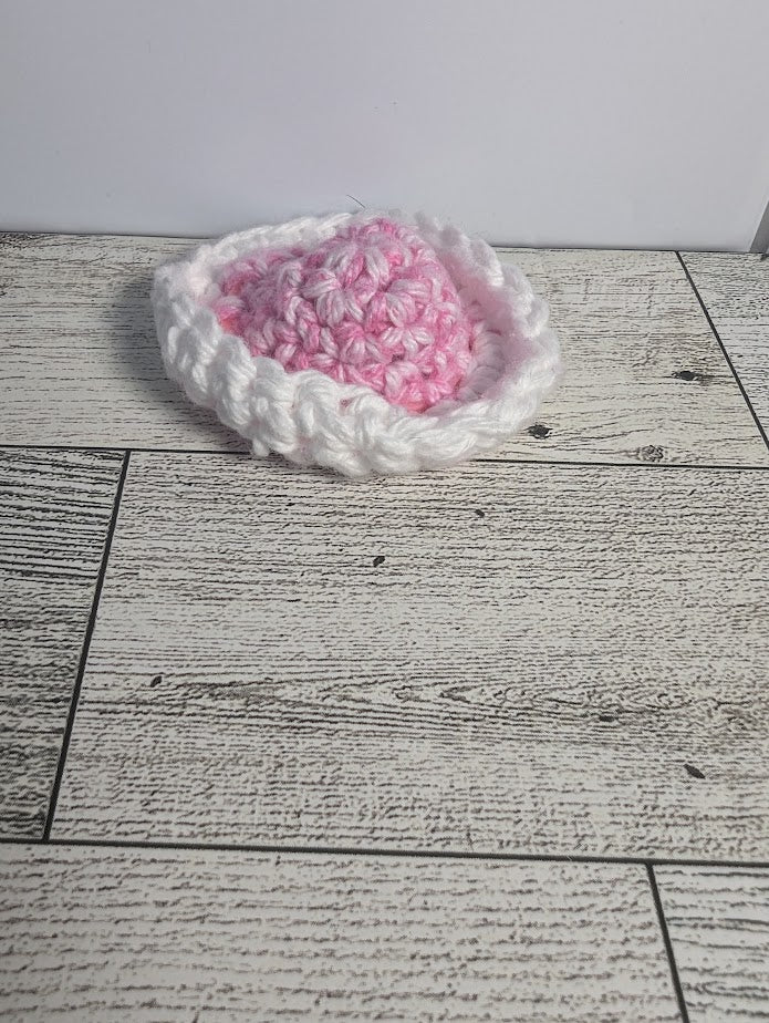 A crochet fidget with a pink center and a white rim. The shape of the fidget resembles a sombrero. The background is a light wood grain and a white tile.