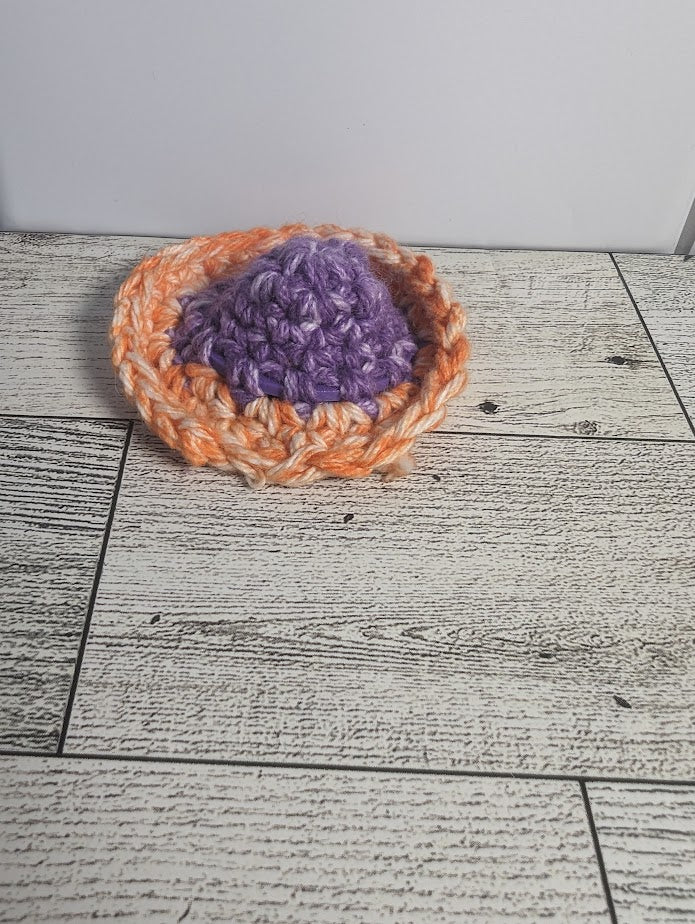 A crochet fidget with a purple center and a orange rim. The shape of the fidget resembles a sombrero. The background is a light wood grain and a white tile.