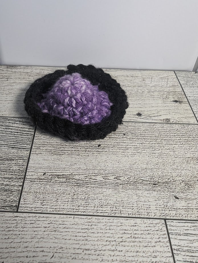 A crochet fidget with a purple center and a black rim. The shape of the fidget resembles a sombrero. The background is a light wood grain and a white tile.