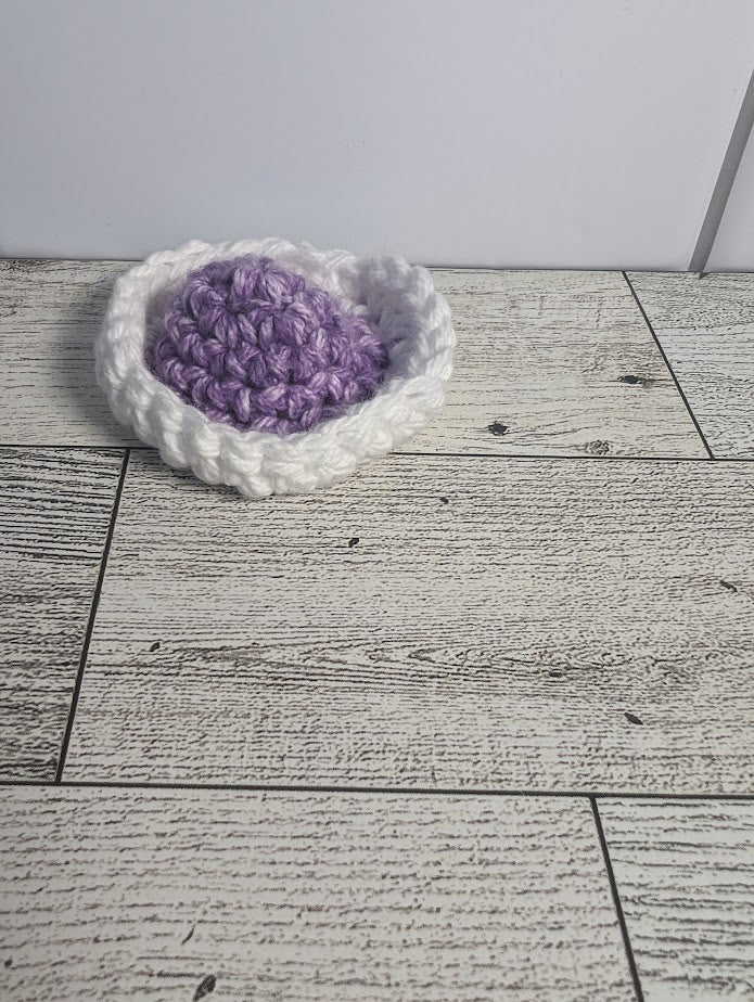 A crochet fidget with a purple center and a white rim. The shape of the fidget resembles a sombrero. The background is a light wood grain and a white tile.