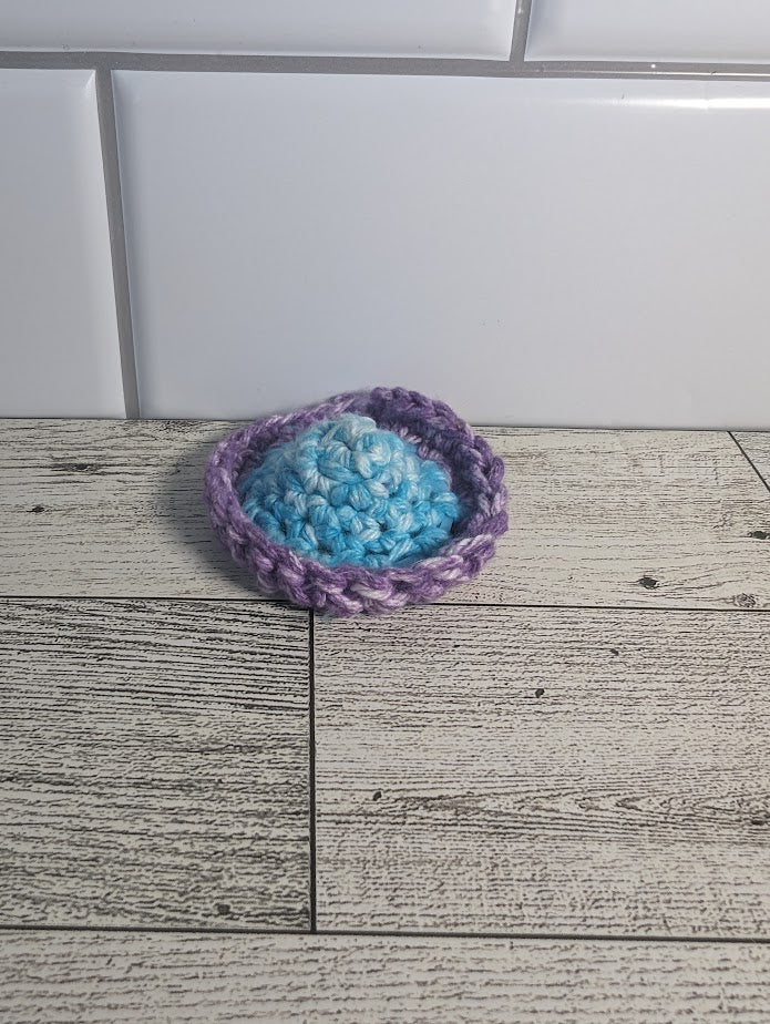A crochet fidget with a light blue center and a purple rim. The shape of the fidget resembles a sombrero. The background is a light wood grain and a white tile.