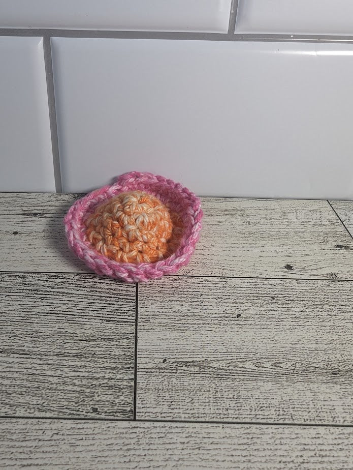 A crochet fidget with an orange center and a pink rim. The shape of the fidget resembles a sombrero. The background is a light wood grain and a white tile.