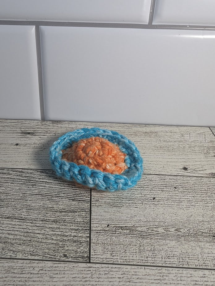 A crochet fidget with an orange center and a light blue rim. The shape of the fidget resembles a sombrero. The background is a light wood grain and a white tile.
