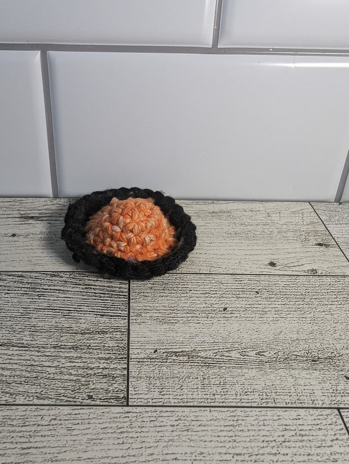 A crochet fidget with an orange center and black rim. The shape of the fidget resembles a sombrero. The background is a light wood grain and a white tile.