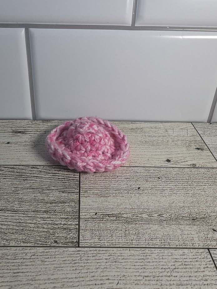 A crochet fidget with both a pink center and a pink rim. The shape of the fidget resembles a sombrero. The background is a light wood grain and a white tile.