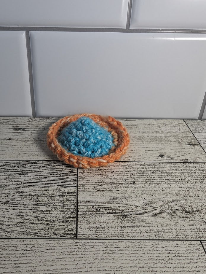 A crochet fidget with a light blue center and orange rim. The shape of the fidget resembles a sombrero. The background is a light wood grain and a white tile.