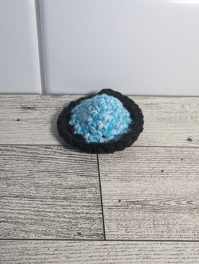 A crochet fidget with a light blue center and a black rim. The shape of the fidget resembles a sombrero. The background is a light wood grain and a white tile.