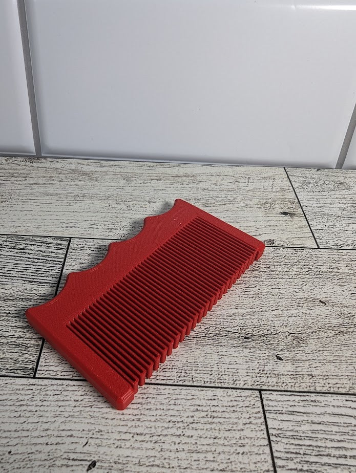 A red comb is on a light wood grain surface. The backdrop is a white tile.