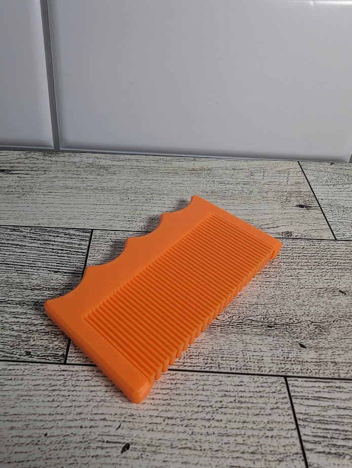 An orange comb is on a light wood grain surface. The backdrop is a white tile