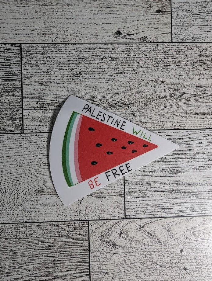 The sticker has the words "Palestine will be free" in the colors black, green, and red. There is a drawn watermelon slice with a green rind, a strip of white, red flesh, and black seeds. The backdrop is a light wood grain.