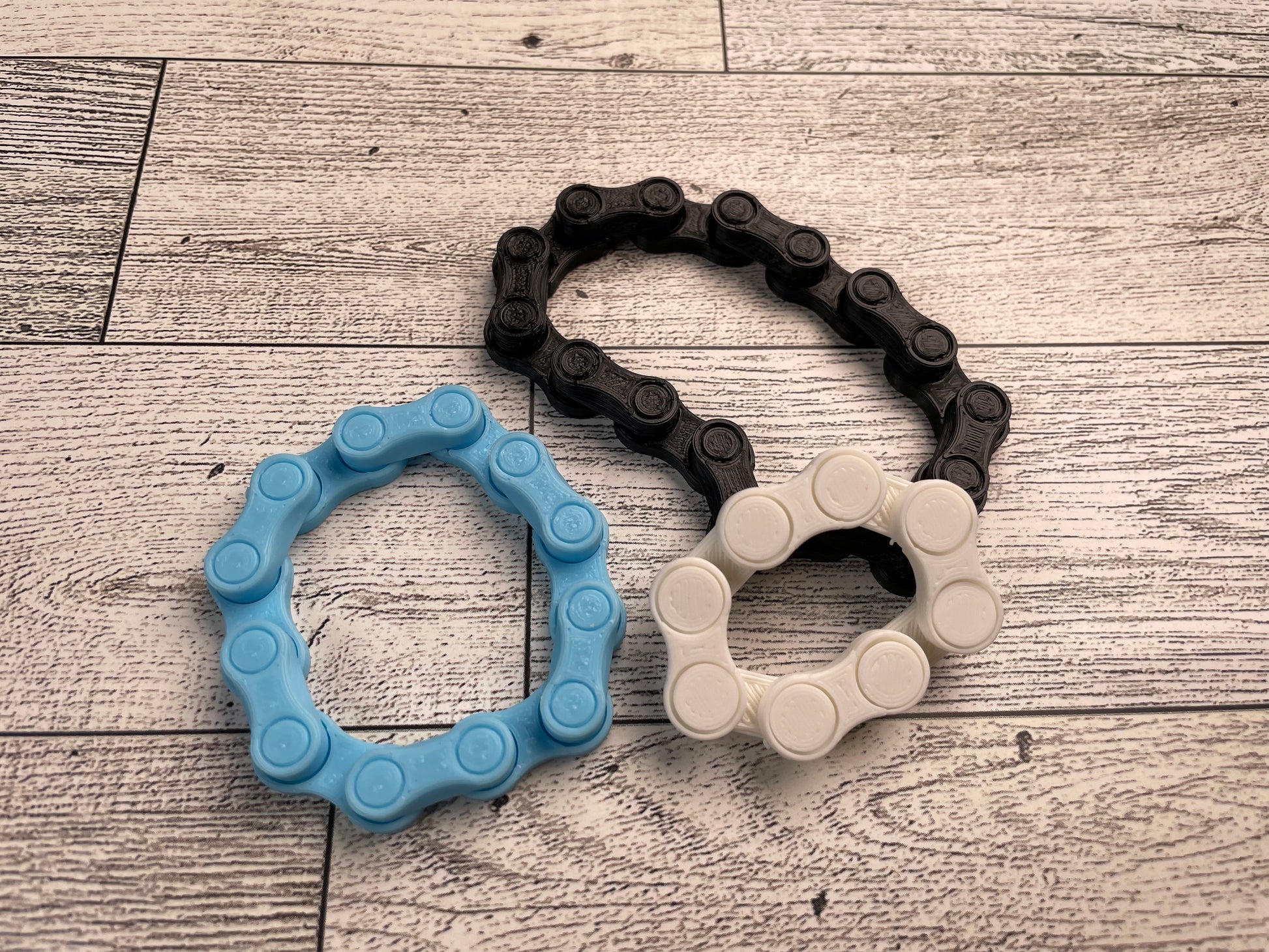 Three chain links in three sizes and colors on a wood backdrop. The bigest chain is black, is arranged in kidney bean shape, and has eight links. Below and to the left of the black chain is a blue chain arranged in a circle and has six links. The last chain is white and is leaning against the bottom of the black chain. This chain is white, has four links and is arranged in a squashed circle.